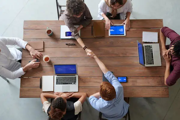 A group of six diverse people in a meeting, shaking hands over a wooden table with laptops, notebooks, and drinks present.