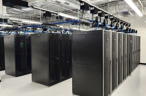 A modern data center featuring rows of black server racks, overhead cable management, and bright fluorescent lighting.