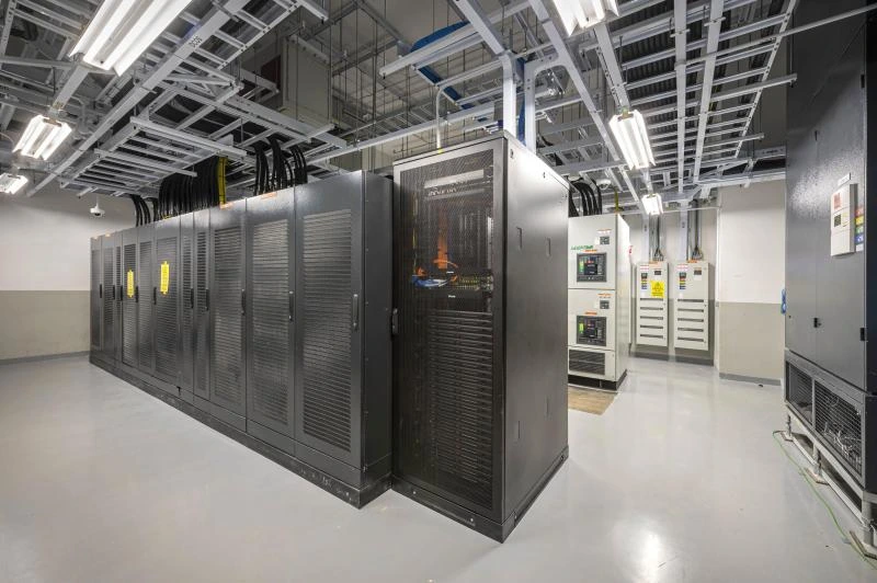 A view of a modern data center with black server racks, bright overhead lighting, and electrical equipment along the walls.
