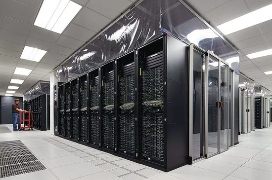 A modern data center with rows of black server racks and a technician working nearby, all under bright fluorescent lighting.