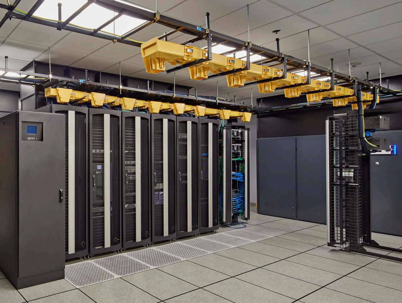 A modern data center featuring tall server racks with cooling systems above, organized cables, and a clean, tiled floor.