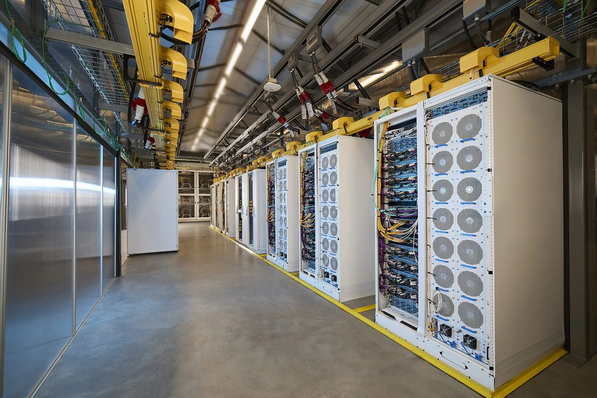 A modern data center aisle featuring rows of server racks with colorful cabling, overhead cable trays, and polished concrete flooring.